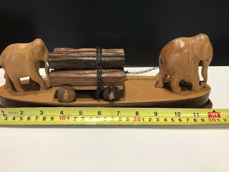 May include: Wooden figurine of two elephants pulling a cart with logs. The cart has wheels and a chain connecting the elephants to the cart. The figurine is on a wooden base.