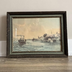 May include: Antique framed print depicting a river scene with a steamboat and other vessels. The artwork features muted tones of blue, green, and beige, set within a dark wooden frame with a green inner border. The print shows a historical landscape.