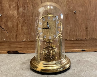 Op de afbeelding: Een goudkleurige antieke klok onder een glazen stolp. De wijzerplaat van de klok heeft Romeinse cijfers en decoratieve details. De klok staat op een ronde voet en heeft een slinger zichtbaar door het glas.