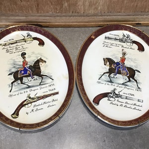 May include: Two white ceramic plates with gold trim, each depicting a soldier on horseback with a pistol. The plates feature text describing the soldier's regiment and the pistol's details. The text on the left plate reads "Officer of the 5th Dragoon Guards, 1833.  circa 1660.  Flintlock Holster Pistol by M. Stones London." The text on the right plate reads "Officer of the 11th Light Dragoons, 1833.  circa 1720.  Cannon Barrel Flintlock Pistol by B. Bronks London."