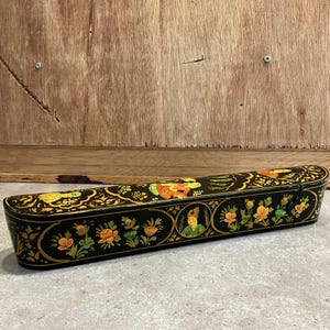 May include: A black, rectangular box with a hinged lid. The box is adorned with intricate floral and figure designs in gold, orange, and green. The box is set against a wooden background.