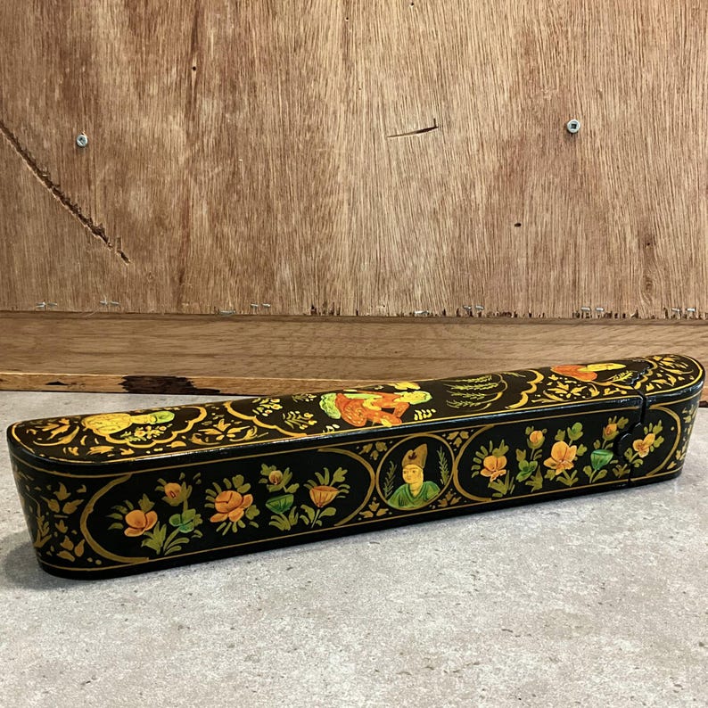 May include: A black, rectangular box with a hinged lid. The box is adorned with intricate floral and figure designs in gold, orange, and green. The box is set against a wooden background.