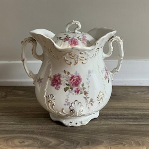 May include: A white porcelain pitcher with a lid, adorned with pink floral patterns and gold accents. The pitcher has two handles and a decorative spout. The floral design includes pink roses and other flowers. The pitcher is a decorative item.