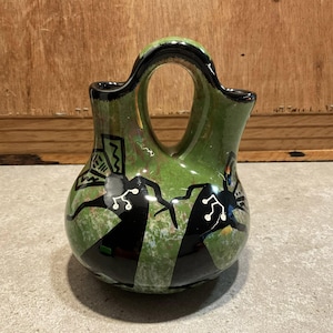 Puede incluir: Un jarrón de cerámica verde con un asa única y dos aberturas. El jarrón presenta diseños abstractos en blanco y negro y un fondo verde moteado. El borde y el asa están pintados de negro. Es un objeto decorativo.