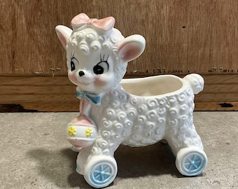 May include: A vintage ceramic lamb planter, white with a textured wool-like design. The lamb has a pink bow, blue accents, and holds a decorative rattle. The planter has blue wheels and is suitable for small plants or decor.