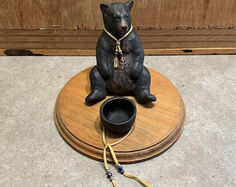 May include: A black bear figurine with a yellow necklace and beaded accents sits on a round wooden base. A small black bowl and a leather cord with beads are in front of the bear. The bear is sitting with its legs spread.