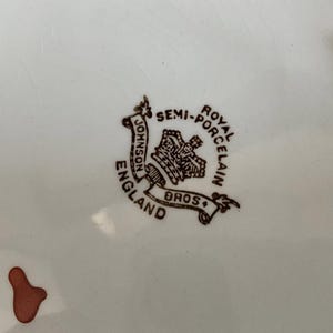 May include: A close-up of a white porcelain plate with a brown emblem. The emblem reads "Johnson Bros England" and "Semi-Royal Porcelain". The plate has a small, brown, abstract shape on the lower left.