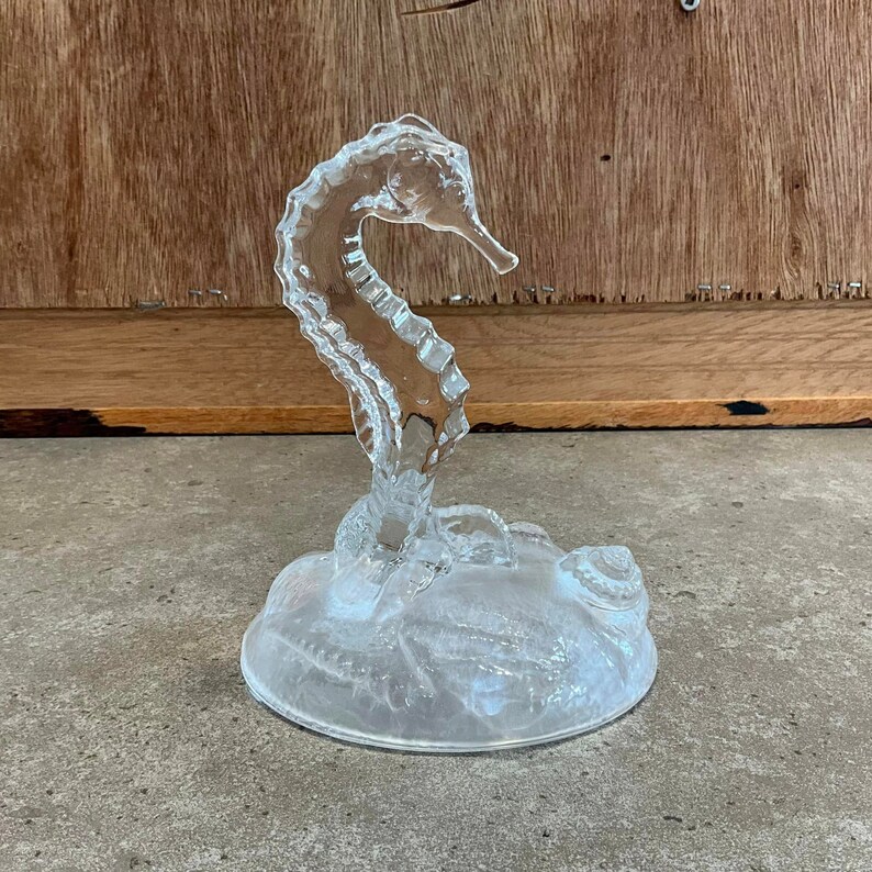 May include: A clear glass seahorse figurine with a detailed design. The seahorse is standing on a base with a textured surface that resembles a seashell.