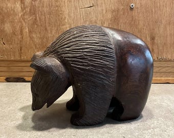 May include: A carved wooden bear figurine in a dark brown color. The bear is depicted in a walking pose, with detailed carving on the fur and body. The figurine is on a gray surface with a wooden background.