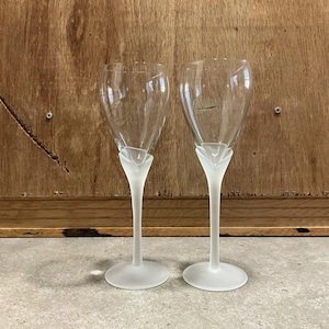May include: Two clear wine glasses with a frosted white base and stem. The bowl of the glass is clear, and the stem has a unique, stylized design. The glasses are set against a wooden background.