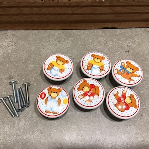 May include: Six white ceramic knobs with a blue and red polka dot pattern. Each knob features a cartoon teddy bear with different outfits and accessories. The knobs are accompanied by six silver screws.