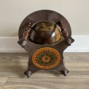 May include: A vintage globe on a wooden stand. The globe is brown with detailed map illustrations. The stand has a compass rose design in orange, green, and yellow, with the text "MADE IN ITALY". The stand and globe are made of wood.
