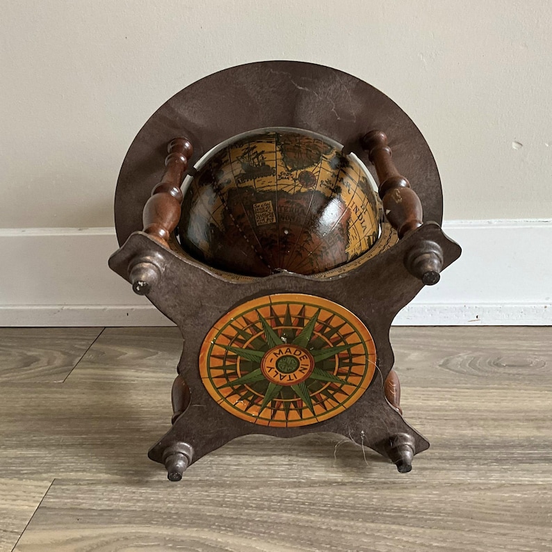 May include: A vintage globe on a wooden stand. The globe is brown with detailed map illustrations. The stand has a compass rose design in orange, green, and yellow, with the text "MADE IN ITALY". The stand and globe are made of wood.
