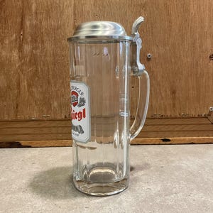 May include: A clear glass beer stein with a hinged metal lid and handle. The stein has a rectangular shape with vertical ridges and a label that reads "Burger Liegl Bier". The stein is designed for holding beer.