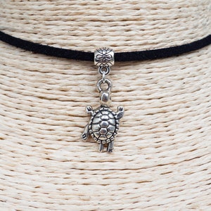 Può includere: Una collana girocollo in velluto nero con un ciondolo a forma di tartaruga color argento. Il ciondolo a forma di tartaruga è dettagliato con un guscio strutturato e piccole zampe. La collana è esposta su un manichino.