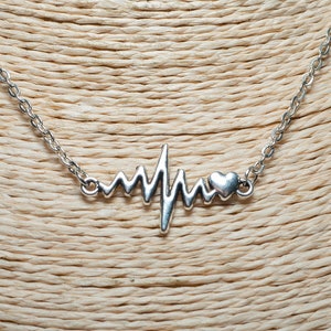 Op de afbeelding: Een zilverkleurige ketting met een hartslagontwerp en een kleine hartvormige bedel. De ketting wordt gepresenteerd op een mannequin in neutrale tinten, waarbij de fijne ketting en hanger worden benadrukt.