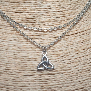 Könnte beinhalten: Halskette mit silberfarbenen, mehrschichtigen Ketten und einem keltischen Knoten-Anhänger. Die Halskette wird auf einer beigefarbenen Schaufensterpuppe präsentiert. Der Anhänger ist ein stilisiertes, dreilappiges Design.