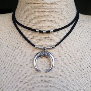 Può includere: Collana con pendente a mezzaluna argentata con cordino nero. La collana ha un design a strati, con un cordino più corto con perline d'argento. Il pendente è dettagliato con motivi intricati, che mostrano uno stile bohémien.
