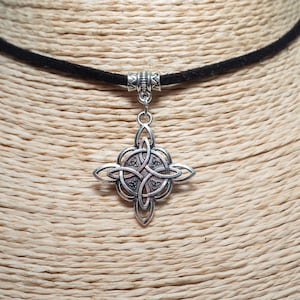 Peut inclure: Un collier pendentif argenté avec un motif de nœud celtique. Le pendentif est suspendu à un choker en velours noir. Le collier est présenté sur un buste de mannequin beige, mettant en valeur le design et la texture du bijou.