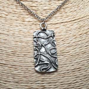 Könnte beinhalten: Eine silberfarbene Halskette mit Anhänger. Der rechteckige Anhänger hat ein abstraktes Design aus wirbelnden Linien. Die Halskette hat eine silberfarbene Kette mit einem filigranen Gliedermuster. Die Halskette wird auf einer neutralfarbenen Schaufensterpuppe präsentiert.