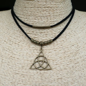 Könnte beinhalten: Eine schwarze Wildleder-Halskette mit einem bronzefarbenen keltischen Knotenanhänger. Die Halskette hat ein doppelreihiges Design mit bronzefarbenen Perlen und einem zylindrischen Akzent. Der Anhänger ist ein detaillierter, dreiteiliger Knoten.