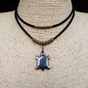 May include: A layered necklace with a dark blue turtle pendant. The pendant hangs from a black cord necklace with bronze-coloured metal accents. The turtle pendant has a textured surface.