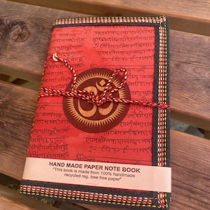 May include: A rectangular notebook with a red cover featuring black Sanskrit script and a central circular emblem with an Om symbol. The notebook is bound with a red, black, and white cord and includes a paper band labelled HAND MADE PAPER NOTE BOOK.