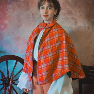 May include: A bright orange plaid cape featuring a rounded collar and a single button closure at the neck. The cape is worn over a white long-sleeved shirt with voluminous, gathered cuffs and solid light orange trousers, creating a layered look.