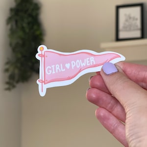 May include: Pink banner with the text "GIRL ♥ POWER" in white lettering. The banner is attached to a white stick with a yellow top.