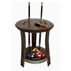 May include: A wooden pool cue rack made from a repurposed barrel with a round top and three legs. The rack has a built-in triangle for holding pool balls and a brush for cleaning the cues. The rack is stained a dark brown color and has a silver metal band around the bottom.