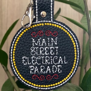 Könnte beinhalten: Ein schwarzer Leder-Schlüsselanhänger mit dem Text "MAIN STREET ELECTRICAL PARADE" in Weiß gestickt. Der Schlüsselanhänger hat einen gelben und weißen Rand und einen silbernen Verschluss.