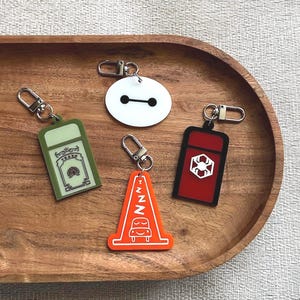 May include: A collection of keychains with various designs, including a white circle with black eyes, a green trash can, a red rectangle with a white symbol, and an orange traffic cone with a car. Each keychain has a silver clasp.
