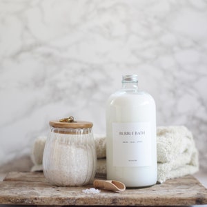 Puede incluir: Un tarro de vidrio transparente con tapa de madera lleno de sales de baño blancas, una botella de vidrio transparente con una etiqueta blanca que dice "BUBBLE BATH" y una pala de madera sobre una superficie de madera.