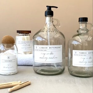 Puede incluir: Una colección de recipientes de vidrio transparente para productos de lavandería con tapas y etiquetas negras. Incluye un dispensador de detergente líquido, una botella de suavizante y frascos para potenciadores de aroma y hojas atrapa-color. Pinzas de ropa de madera en primer plano.