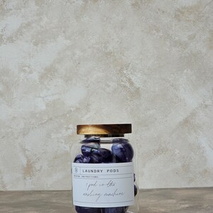 May include: A glass jar with a wooden lid filled with purple laundry pods. The label on the jar reads "Laundry Pods" and "Washing Instructions: 1 pod in the washing machine."