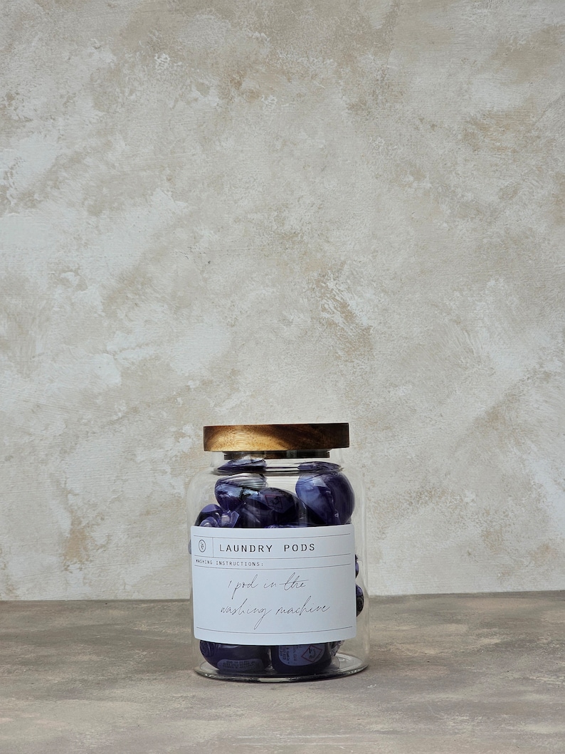 May include: A glass jar with a wooden lid filled with purple laundry pods. The label on the jar reads "Laundry Pods" and "Washing Instructions: 1 pod in the washing machine."