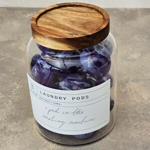 May include: A glass jar with a wooden lid filled with purple laundry pods. The label on the jar reads "Laundry Pods" and "1 pod in the washing machine".