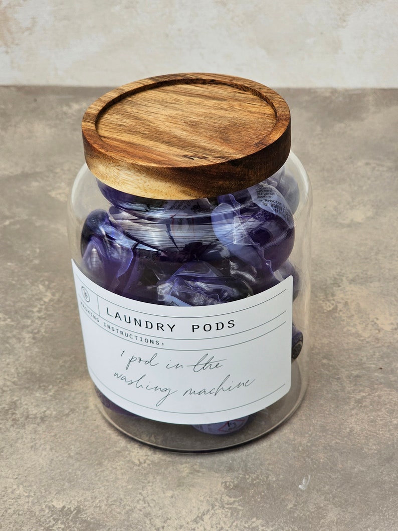 May include: A glass jar with a wooden lid filled with purple laundry pods. The label on the jar reads "Laundry Pods" and "1 pod in the washing machine".