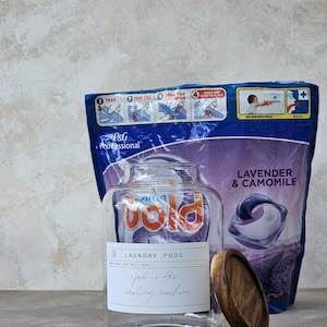 May include: A clear glass jar with a wooden lid filled with laundry detergent pods. The jar has a label that says "Laundry Pods" and "1 pod in the washing machine". The jar is sitting on a light grey surface in front of a purple and blue bag of laundry detergent pods. The bag has the words "Lavender & Camomile" printed on it.