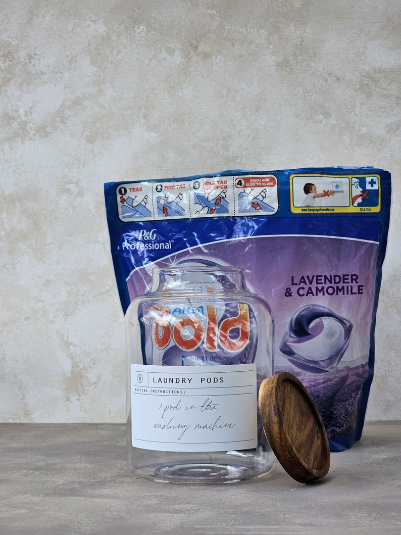 May include: A clear glass jar with a wooden lid filled with laundry detergent pods. The jar has a label that says "Laundry Pods" and "1 pod in the washing machine". The jar is sitting on a light grey surface in front of a purple and blue bag of laundry detergent pods. The bag has the words "Lavender & Camomile" printed on it.