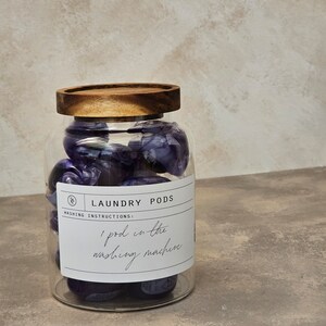 May include: A glass jar with a wooden lid filled with purple laundry detergent pods. The label on the jar reads "Laundry Pods" and "Washing Instructions: 1 pod in the washing machine."