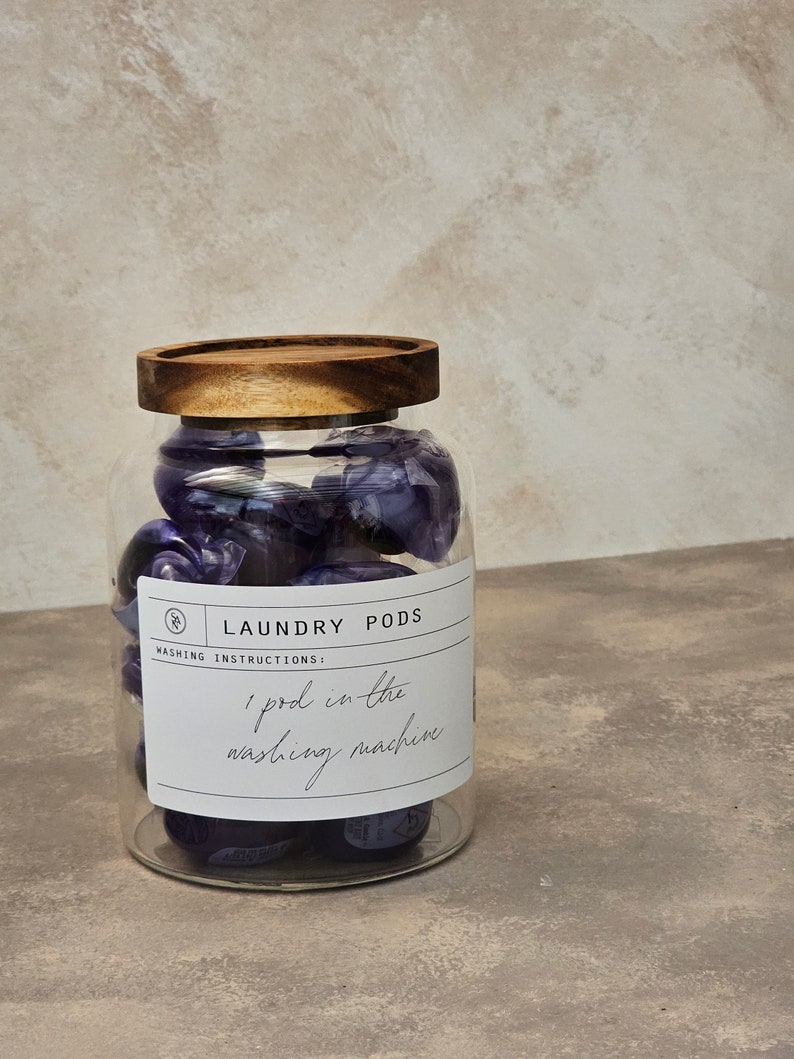 May include: A glass jar with a wooden lid filled with purple laundry detergent pods. The label on the jar reads "Laundry Pods" and "Washing Instructions: 1 pod in the washing machine."