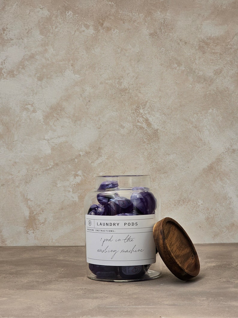 May include: A glass jar filled with purple laundry detergent pods. The jar has a wooden lid and a label that reads "Laundry Pods" and "1 pod in the washing machine".