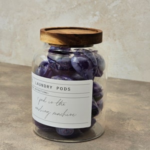 May include: A glass jar with a wooden lid filled with purple laundry detergent pods. The jar has a label that says "Laundry Pods" and "1 pod in the washing machine".
