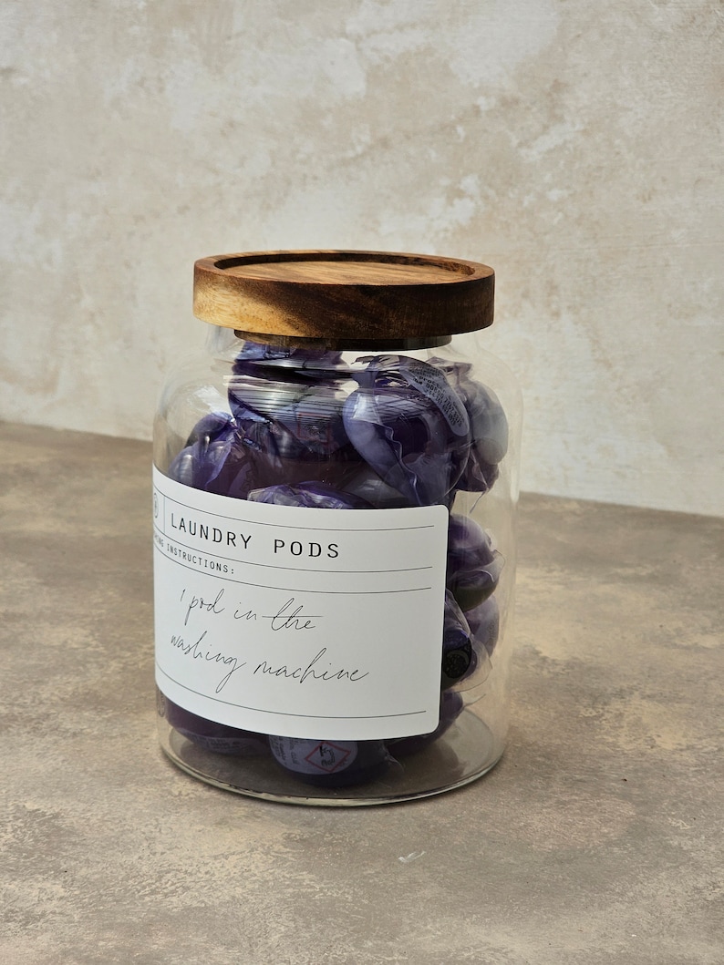 May include: A glass jar with a wooden lid filled with purple laundry detergent pods. The jar has a label that says "Laundry Pods" and "1 pod in the washing machine".
