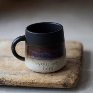 May include: A ceramic mug with a black rim and handle, transitioning to brown and purple hues, and a cream-coloured base. The text "#your text here" is etched on the base. The mug sits on a wooden surface.