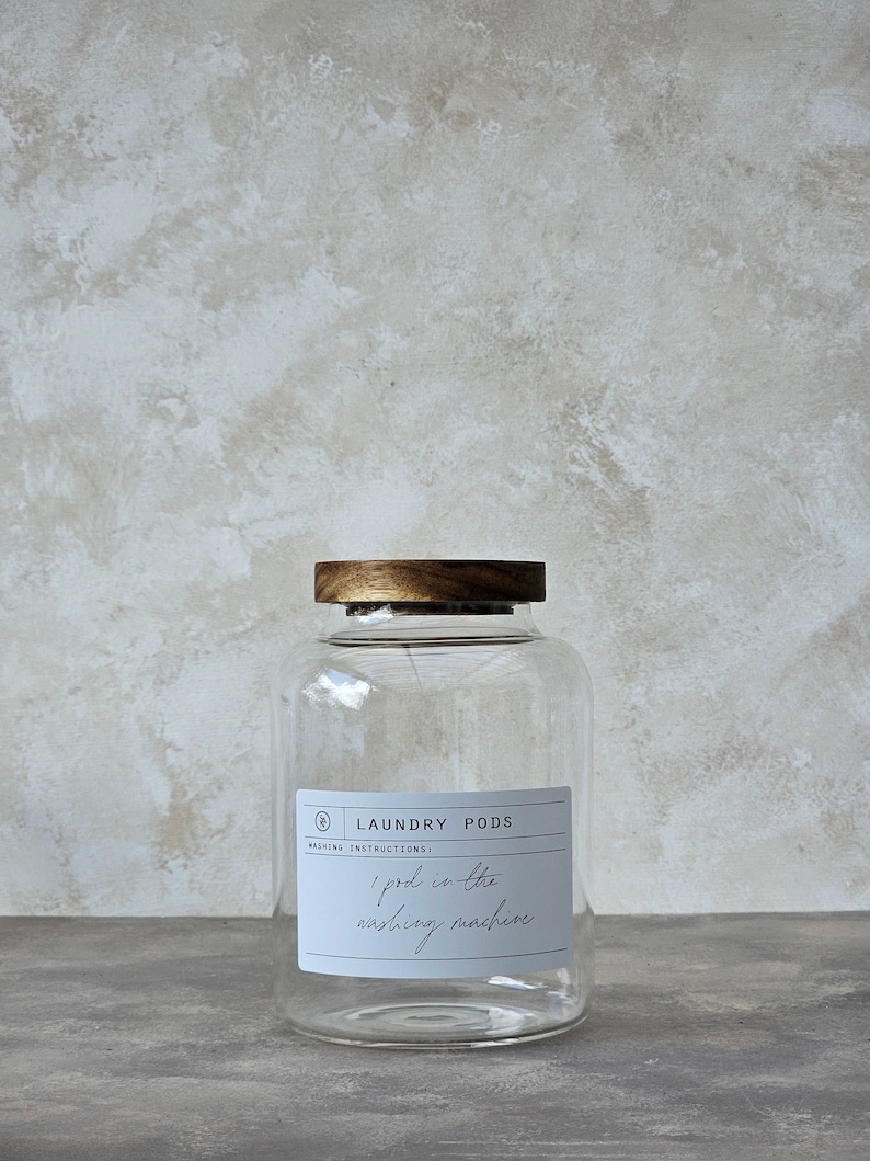 May include: A clear glass jar with a wooden lid, labelled "Laundry Pods". The jar has a white label with the text "Washing Instructions: 1 pod in the washing machine".