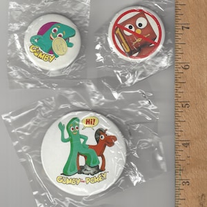 May include: A collection of vintage Gumby and Pokey pinback buttons. The buttons feature colorful illustrations of the characters, including Gumby with a coin, Gumby and Pokey together, and Pokey alone. The buttons are packaged in clear plastic.