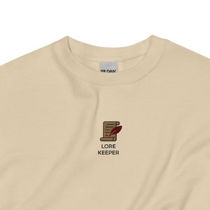 May include: A cream-colored sweatshirt with the text "LORE KEEPER" and a small graphic of a scroll and quill. The sweatshirt is made of a soft material and has a crew neck and long sleeves. The Gildan logo is visible on the upper back.