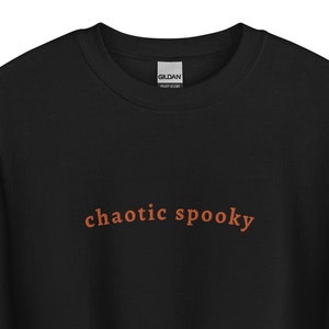 May include: A black long-sleeve crewneck sweatshirt featuring the text "chaotic spooky" printed in a burnt orange color across the center of the chest. The garment has a ribbed collar, cuffs, and hem, with a Gildan brand label at the neckline.